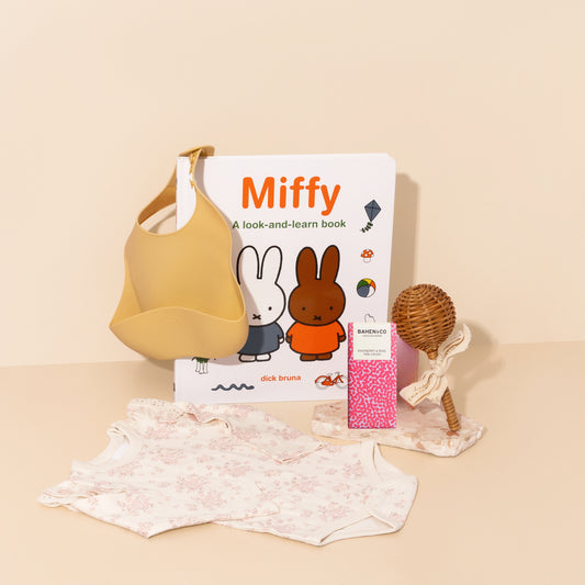 newborn baby girl gift hamper filled with baby essentials such as onesie , silicone bib , rattan rattle , story book and chocolate treat